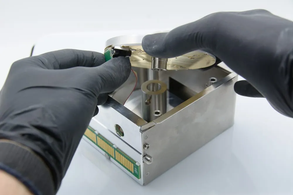 Gloved hands lowering the gold flex-circuit sample carrier disc into the open shuttle chassis.