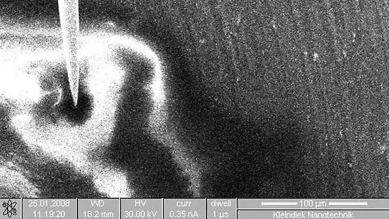 SEM image of a probe tip delivering a splash of glue onto a sample surface, with a 100 micrometre scale bar.