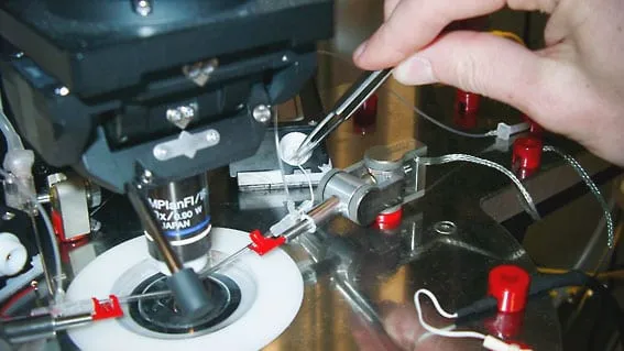 A hand loading a glass pipette into an MM3A-LS pipette holder beside a microscope objective.