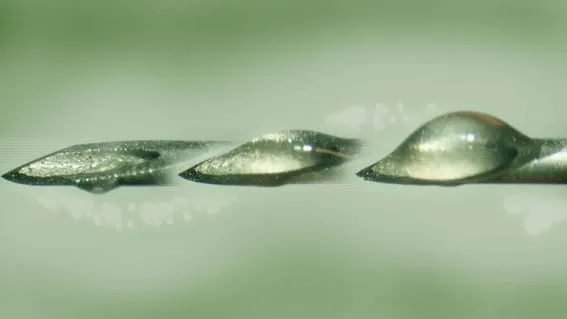 Macro photograph of three glistening SemGlu adhesive droplets of increasing size on a tip against a green background.