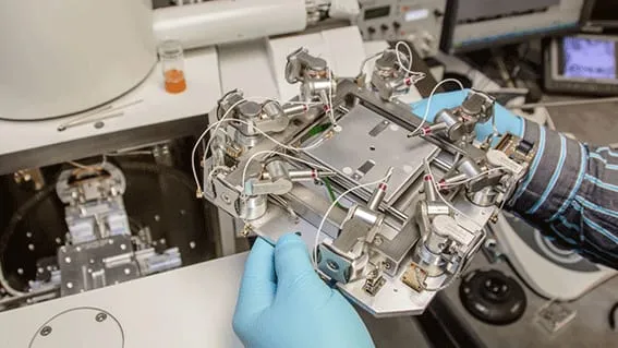 Gloved hands carrying the complete ProbeWorkstation+ platform towards an open SEM chamber.